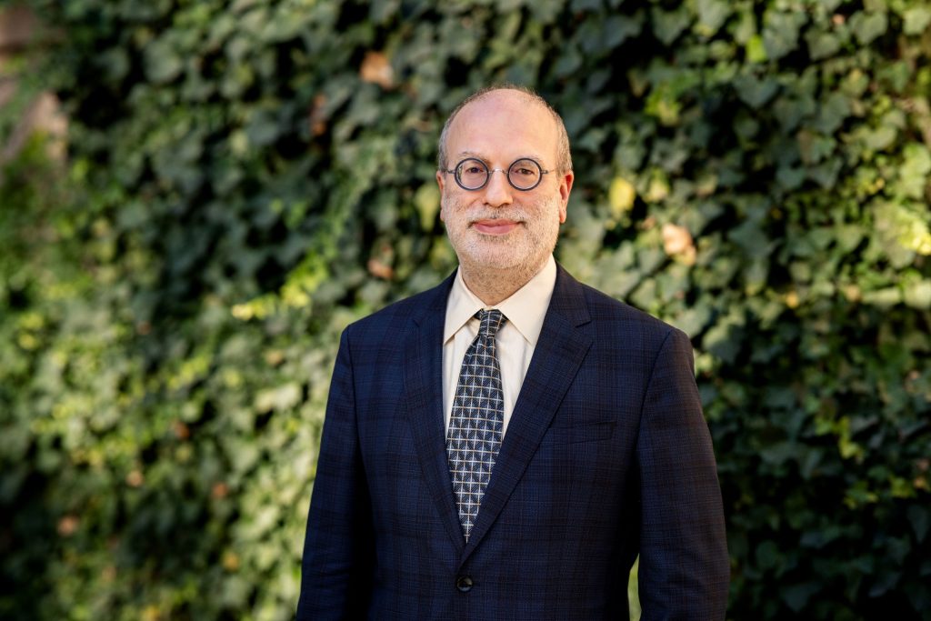 Portrait of guest speaker Dr. Michael Gordin. The subject is staring at the camera. He is wearing a blue plaid suit and tie with geometric shapes. Behind the subject is a building covered in green ivy.