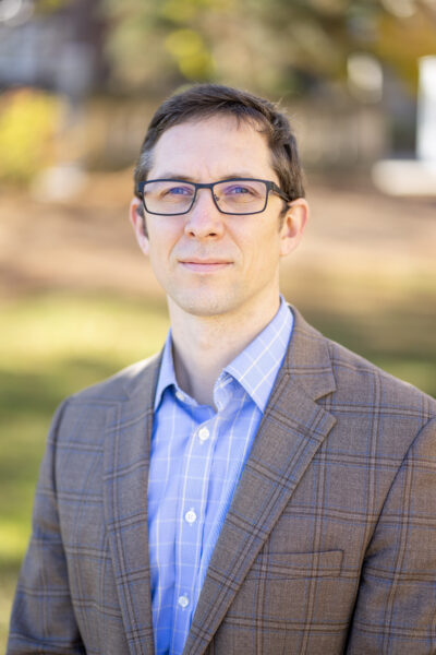 Headshot photograph of Professor Patrick Iber. Iber looks at the camera, wearing glasses and a grey suit, with grass and leaves in the background.